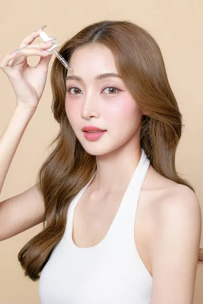若いアジアの美女は 顔に韓国のメイクスタイルとピペットを使用して 孤立したベージュの背景に皮膚に血清を適用するカーリー長い髪 フェイシャルトリートメント プラスチック手術 — ストック写真
