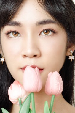 Kakülleri ve pürüzsüz cildi olan güzel, genç Asyalı kadın yüzüne yakın çekim. Doğal makyajlı, beyaz arka planda çiçek tutan güzel bir bayan model. Kozmetik, güzellik ve kaplıca.