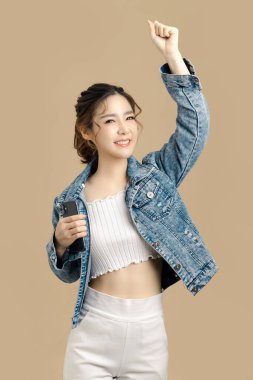 Gülümse güzel Asyalı kadın bir cep telefonu tutuyor ve kollarını kaldırıyor başarısını tebrik ediyor. Kameraya bak, kopyala alanı, izole edilmiş bej arkaplan.