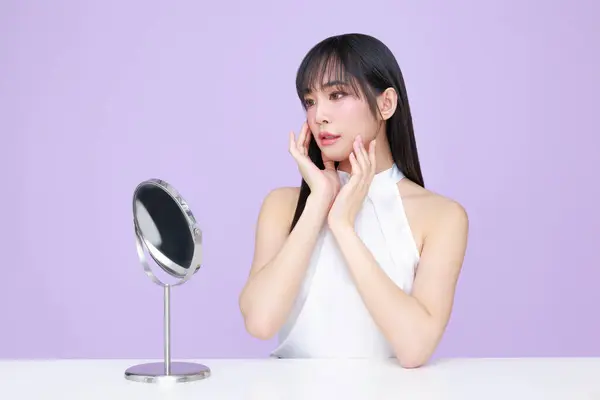 Beautiful young Asian woman model long hair with natural makeup on face clean skin with mirror on isolated violet background. Cute girl portrait, Facial treatment, Body care, Beauty and Spa.