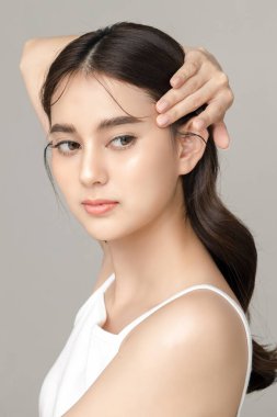 Güzel, genç, Asyalı bir manken, gri arka planda tertemiz bir ciltle yüzüne dokunuyor. Yüz bakımı, yüz bakımı, kozmetik, estetik cerrahi stüdyoda güzel bir kız portresi..