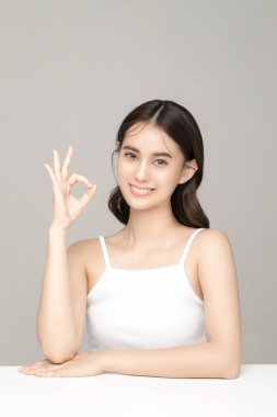 Güzel, genç, Asyalı bir kadın model. Gri arka planda tertemiz bir cilde sahip, tamam işareti gösteriyor. Yüz bakımı, yüz bakımı, kozmetik, estetik cerrahi stüdyoda güzel bir kız portresi..
