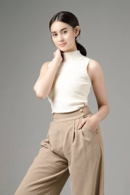 Kendine güvenen Asyalı kadın ellerini cebine koyar ve gri arkaplanda izole edilmiş kameraya bakar. Stüdyoda güzel bir kızın portresi..