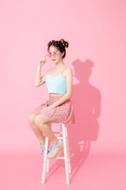 Mavi, şirin, mutlu Asyalı bir kadının portresi güneş gözlüğü takıyor, gülümsüyor ve izole edilmiş pastel pembe arka planda poz veriyor..