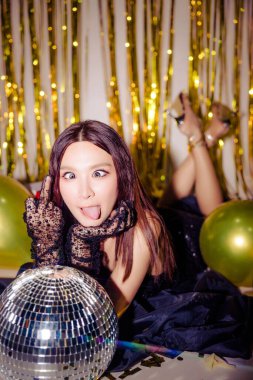 Seksi siyah elbise giyen genç çekici Asyalı kadın balonlarla birlikte parlak altın yaldızlı arka planda parti orta parmağını kutluyor. doğum günü, yeni yıl konsepti.