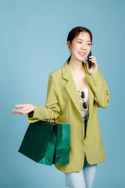 Gayri resmi moda ceketli, güneş gözlüklü, elinde alışveriş çantaları olan, pastel mavi arka planda izole edilmiş cep telefonu kullanan güzel, çekici bir Asyalı kadın. Kadın tüketimi, yaşam tarzı konsepti.