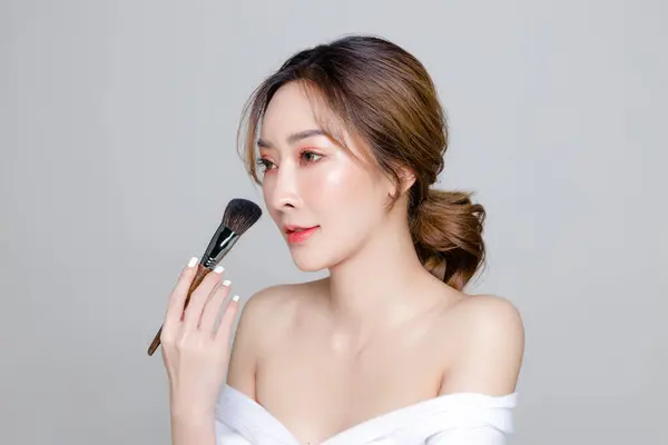 Asian woman with a beautiful face and Perfect clean fresh skin. Portrait of female model using brush for makeup on Grey isolated background. Cosmetology, Body care, plastic surgery, beauty and spa.