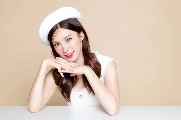 Cute Asian woman with perfect clear fresh skin. Pretty girl model wearing white beret and natural makeup on beige background. Cosmetology, beauty and spa, wellness, Plastic surgery.