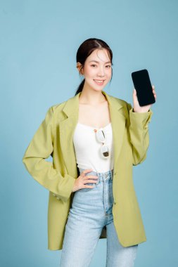Gayri resmi yeşil moda ceketli ve güneş gözlüklü genç ve çekici Asyalı kadın pastel mavi arka planda izole edilmiş boş ekran cep telefonu gösteriyor. Kadın tüketimi, yaşam tarzı konsepti.