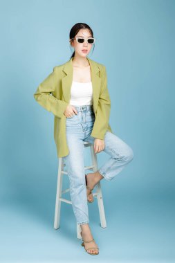 Yeşil moda ceketli, güneş gözlüklü, pastel mavi arka planda izole edilmiş bir sandalyede oturan güzel ve çekici bir Asyalı kadın. Kadın tüketimi, yaşam tarzı konsepti.