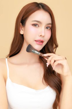 Güzel, genç, Asyalı bir kadın, uzun saçlı, yüzüne makyaj yapmak için fırça kullanıyor. İzole edilmiş bej arka planda temiz bir ten. Tatlı kız portresi, yüz bakımı, vücut bakımı, güzellik ve güzellik merkezi..