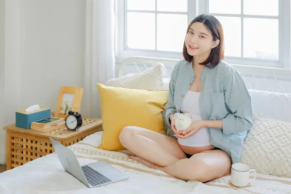 若いアジアの幸せな妊娠中の女性は ベッドの上にピギーバンクを保持し 彼女の腹に触れるノートパソコンを使用しています ファミリープラン 期待のコンセプト — ストック写真