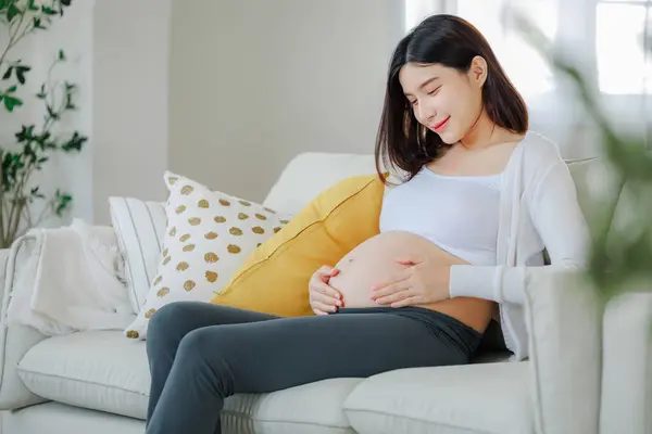 若いアジアの幸せな妊婦がソファーに座って腹に触れています ファミリープラン 子供と母性ケアのコンセプト — ストック写真