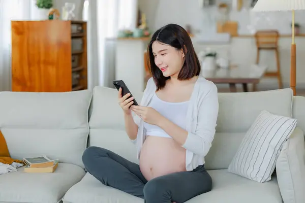 Mladá Asijská Šťastná Těhotná Žena Sedí Pomocí Mobilního Telefonu Pohovce — Stock fotografie