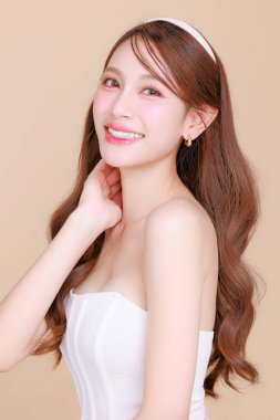 Güzel Asyalı, kıvırcık saçlı, uzun kıvırcık saçlı, Koreli makyajlı, yüzü parlayan ve bej arkaplanda sağlıklı bir yüz portresi olan bir kadın. Kozmetik, estetik cerrahi.