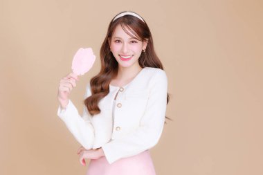 Güzel Asyalı, kıvırcık saçlı, kıvırcık saçlı bir kadın Koreli makyajlı, parlayan bir aynaya ve izole edilmiş bej arka planda sağlıklı bir yüz portresi gülüşüne bakıyor. Kozmetik, estetik cerrahi.