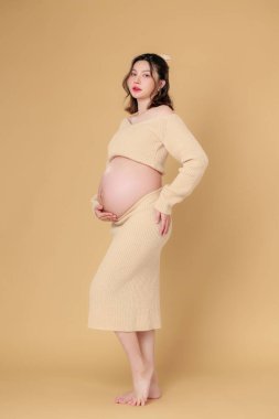 Güzel Asyalı Hamile Mutluluk Kadını örgü gömlekli, göbeğine dokunmuş, bej renkli, soyutlanmış arka planda doğmamış bebeği bekleyen gülümsüyor. Annem karnını tutuyor. Anneliğe yolculuk.