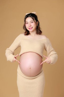 Güzel Asyalı Hamile Mutluluk kadını örgü gömleğiyle gülümseyip göbeğini göstererek bej rengi izole bir arka planda doğmamış bebeği bekliyor. Annem karnını tutuyor. Anneliğe yolculuk.