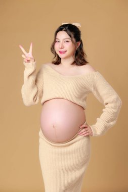 Güzel Asyalı Hamile Mutluluk Kadını örgü gömlekli, göbeğine dokunmuş, bej renkli, soyutlanmış arka planda doğmamış bebeği bekleyen gülümsüyor. Annem karnını tutuyor. Anneliğe yolculuk.