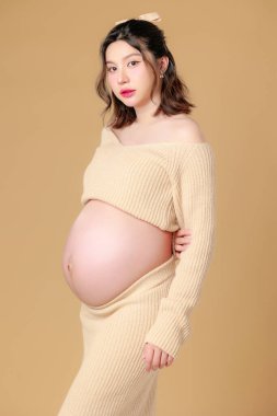 Güzel Asyalı Hamile Mutluluk Kadını örgü gömlekli, göbeğine dokunmuş, bej renkli, soyutlanmış arka planda doğmamış bebeği bekleyen gülümsüyor. Annem karnını tutuyor. Anneliğe yolculuk.