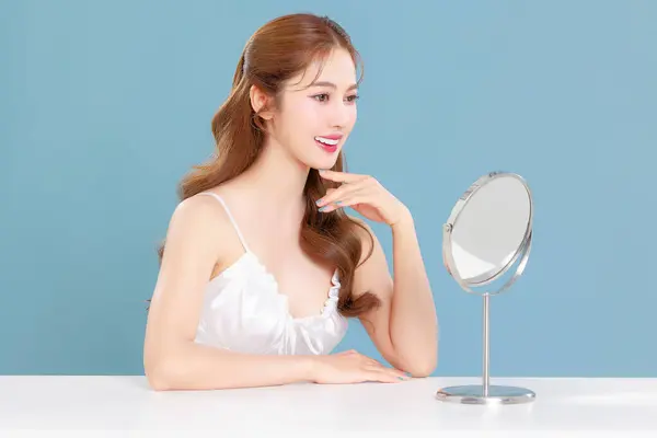 stock image Pretty Asian beauty woman curly long hair with Korean makeup glowing face and healthy facial skin portrait smile and looking at mirror on isolated blue background.