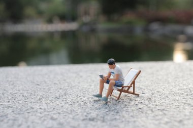 Minyatür bir figür terasta oturuyor, okuyor, huzurlu bir gölün kenarında.