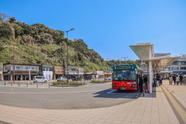 Kırmızı bir otobüse binen yolcuların olduğu bir otobüs durağı. Güneşli bir ortam