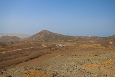 Uzak küçük kulübelerin olduğu çorak çöl manzarası, açık mavi bir gökyüzü.