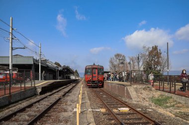 Güneşli bir günde kalabalık bir tren istasyonuna bir tren gelir.,