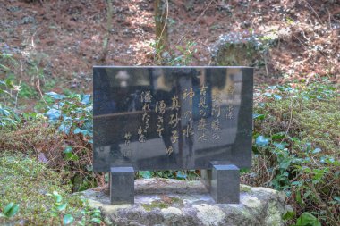 Parlatılmış siyah granit levha, kabartmalı Japon yazısıyla, yemyeşil bitki örtüsüyle çevrili.