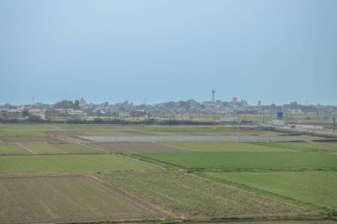 Yeşil tarlalar bulutlu bir gökyüzünün altında uzanır. Uzak bir kasabaya giden bir yol vardır.,