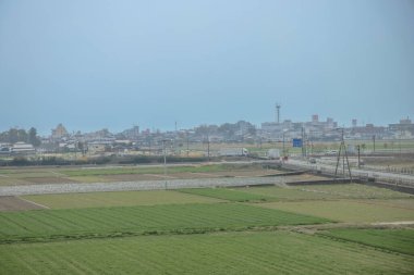 Yeşil tarlalar bulutlu bir gökyüzünün altında uzanır. Uzak bir kasabaya giden bir yol vardır.,