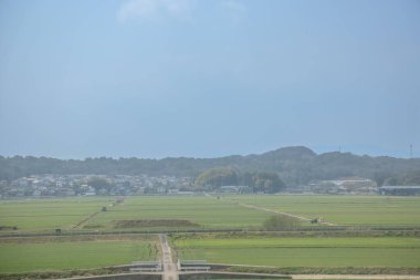 Yeşil tarlalar bulutlu bir gökyüzünün altında uzanır. Uzak bir kasabaya giden bir yol vardır.,