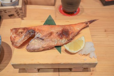 Tahta bir tabakta limon dilimi ve garnitürle sunulmuş ızgara balık.