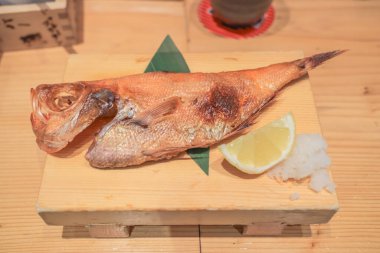 Tahta bir tabakta limon dilimi ve garnitürle sunulmuş ızgara balık.