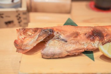 Tahta bir tabakta limon dilimi ve garnitürle sunulmuş ızgara balık.