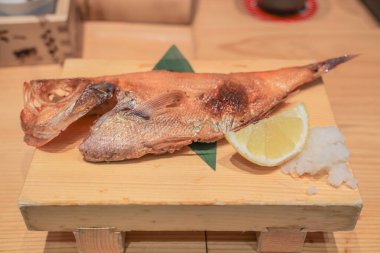 Tahta bir tabakta limon dilimi ve garnitürle sunulmuş ızgara balık.