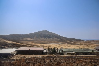 Uzak bir tepe berrak mavi bir gökyüzünün altında yükseliyor, dağınık yapılarla.