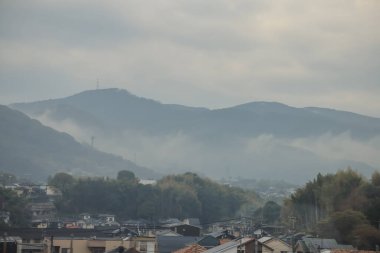 Bulutlu bir gökyüzünün altında sakin bir yerleşim bölgesinin arkasındaki tepeler sakinliği ve doğallığı çağrıştırıyor.