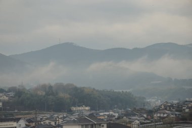 Bulutlu bir gökyüzünün altında sakin bir yerleşim bölgesinin arkasındaki tepeler sakinliği ve doğallığı çağrıştırıyor.