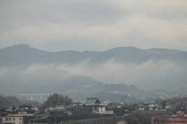 Bulutlu bir gökyüzünün altında sakin bir yerleşim bölgesinin arkasındaki tepeler sakinliği ve doğallığı çağrıştırıyor.