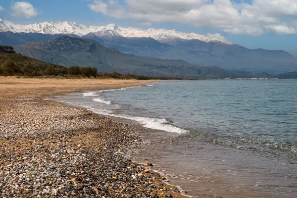 Batı Girit 'te kayalık bir sahil boyunca yumuşak dalgalar kırılıyor; uzak mesafede kar kaplı beyaz dağlar var.