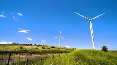 Golan Tepeleri 'ndeki yemyeşil üzüm bağlarının arasında zarif bir şekilde dönen rüzgâr türbinlerinin sakin manzarasını gösteren bir video. Sahne, yenilenebilir enerjiyi tarımla harmanlıyor, engin gökyüzünün altında kayan tepeler ve modern türbinler bulunuyor.. 