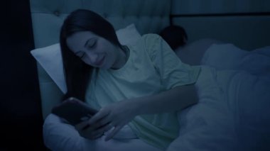 Kocası yatakta uyurken cep telefonu kullanan genç bir kadın.