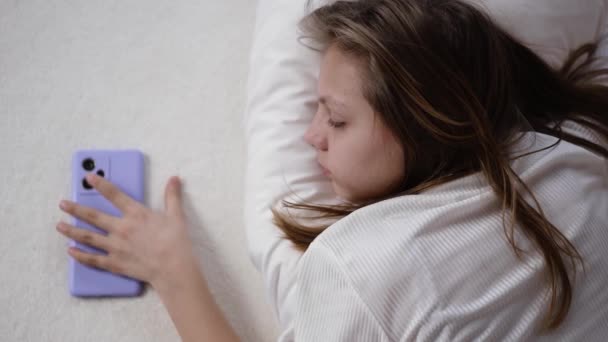 若い女の子が携帯電話の隣のベッドに横たわり 興味を持って画面を見ています — ストック動画
