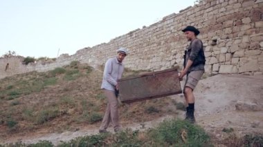 İki adam taş duvarların yanında büyük tahta bir sandık taşıyor..