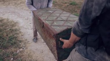 İki adam taş duvarların yanında büyük tahta bir sandık taşıyor..