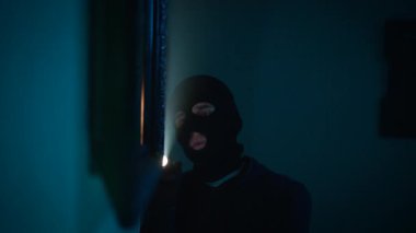 Kar maskeli bir soyguncu bir tablonun arkasında kasa arıyor.