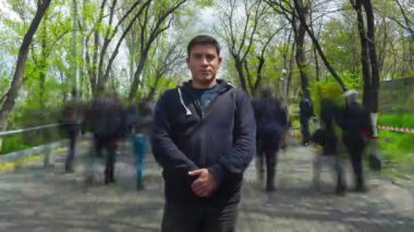 Yalnız bir adam bulanık insanların ve yeşil ağaçların arka planında hareketsiz duruyor. 4K Zamanaşımı videosu.
