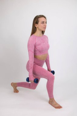 Pembe eşofmanlı bir fitness eğitmeni halterli ısınma egzersizleri gösterir. Işık arkaplanına yakın plan.